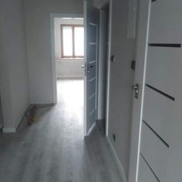 LukBud - Szare panele podłogowe w korytarzu z trzema białymi drzwiami z ciemnymi wstawkami, widok na okno w oddali.