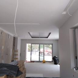 LukBud - Wnętrze w trakcie remontu: ściany po malowaniu na szaro, widoczne kable wystające z sufitu, okno przesuwne z widokiem na zewnątrz, podłoga zabezpieczona folią, kartony z drzwiami i drabina w rogu.