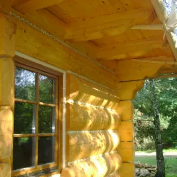 Fragment ściany domu z bali z oknem, widoczne uszczelnienia między balami, ozdobne wykończenie dachu z naturalnego drewna, fragment drzewa w tle.