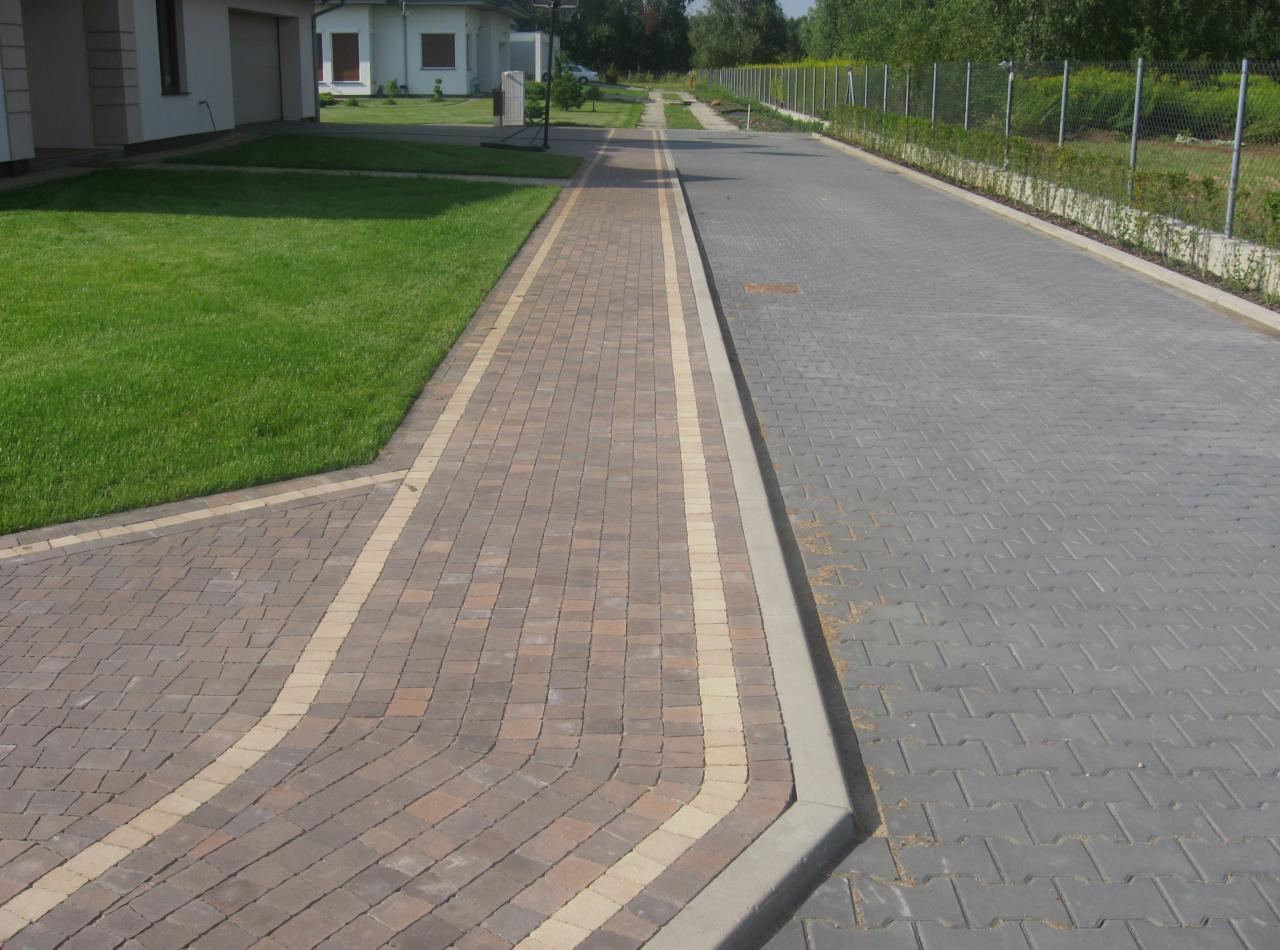 Ułożona ścieżka z kostki brukowej w odcieniach brązu i beżu, oddzielona od szarej nawierzchni betonowym obrzeżem, przylegająca do zadbanego trawnika i ogrodzenia posesji.
