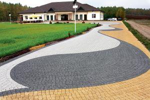 Zakrzywiony podjazd z kostki brukowej w trzech kolorach (szary, biały, żółty) prowadzący do dużego domu z trawnikiem. Widoczna lampa oświetleniowa i samochód w oddali.