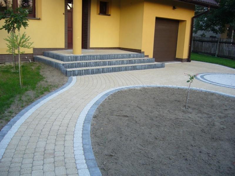 Żółty dom z brukowanym wejściem i ścieżką z jasnej kostki, obrzeża z szarej kostki, świeżo posadzony krzew róży na pierwszym planie.