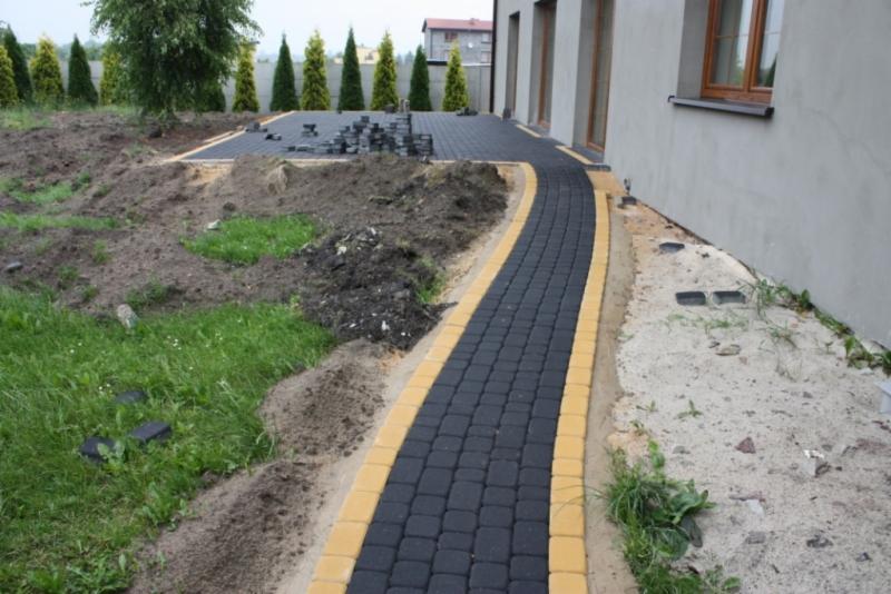 Układanie czarnej kostki brukowej z żółtymi obrzeżami wzdłuż ścieżki i na tarasie, częściowo ułożona, stos kostki obok, przygotowany teren, widok na dom i tuje.