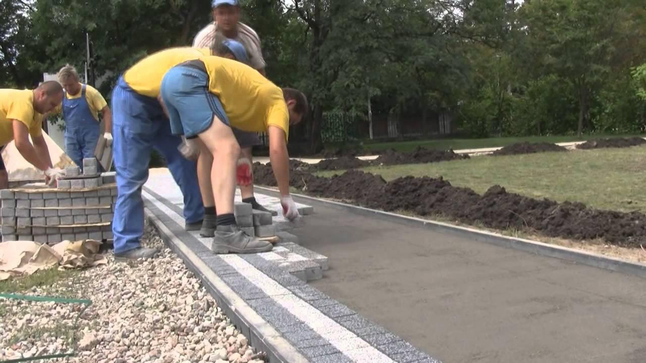 Robotnicy w żółtych koszulkach układają szaro-białą kostkę na przygotowanym podłożu, widoczne stosy kostki i tło z zielenią.