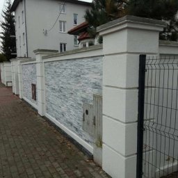 LUX DOM Dmytro Morykot - Nowoczesny, jasnoszary mur z dekoracyjną okładziną z kamienia elewacyjnego, zakończony białymi słupkami z betonowych bloków, sąsiadujący z metalową siatką ogrodzeniową i chodnikiem z kostki brukowej.