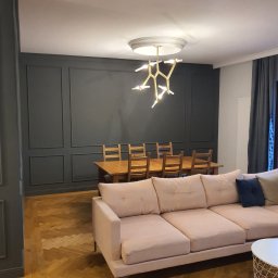 LUX DOM Dmytro Morykot - Salon z ciemnoszarą ścianą z dekoracyjnymi panelami, drewnianym stołem i krzesłami oraz różową sofą, oświetlony nowoczesną lampą sufitową.