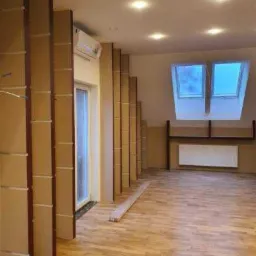 Poddasze z drewnianą podłogą, oknami dachowymi i konstrukcją z płyt kartonowo-gipsowych w trakcie montażu, widoczny klimatyzator na ścianie.