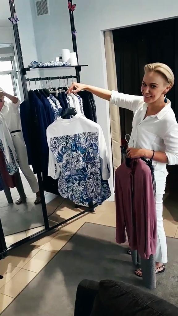 Kobieta w białym komplecie prezentuje koszulę z niebieskim, roślinnym wzorem na plecach oraz bordową bluzkę na wieszaku w butiku.