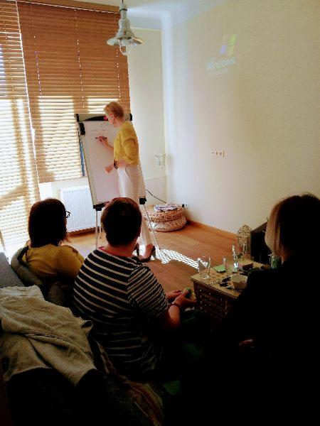 Kobieta w żółtej bluzce pisze na flipcharcie podczas spotkania z grupą osób siedzących na kanapie; na ścianie wyświetlany jest obraz z projektora.