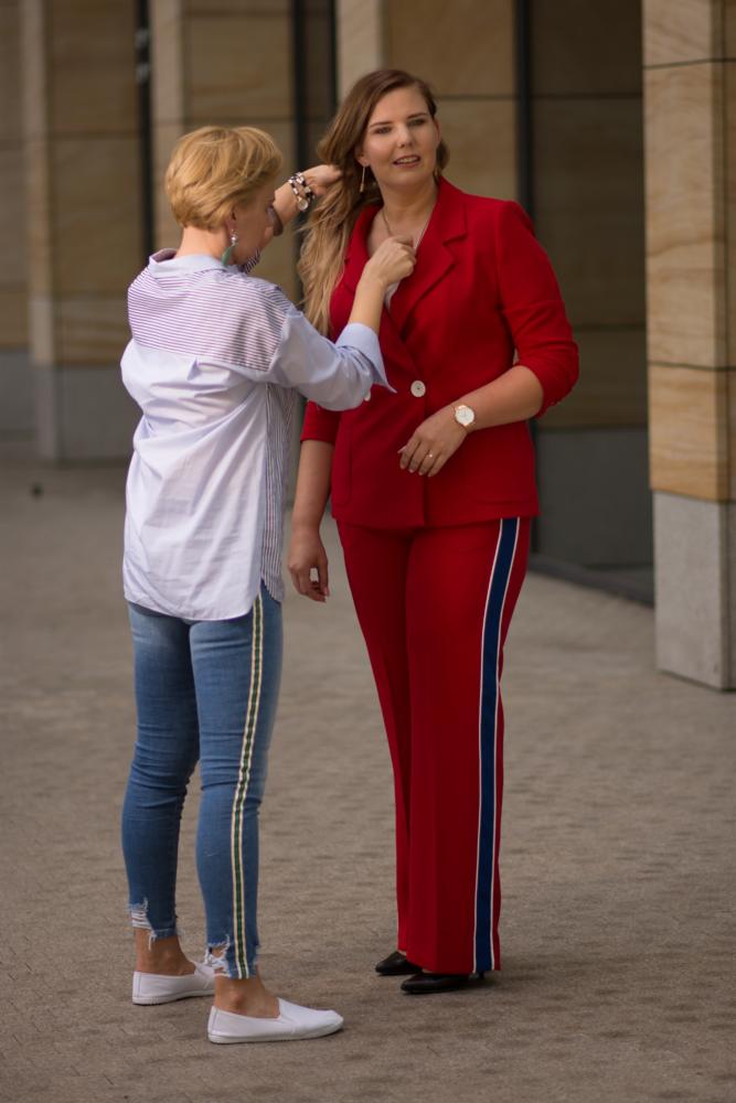 Stylistka poprawia włosy klientce w czerwonym garniturze z lampasem, na tle nowoczesnej architektury.