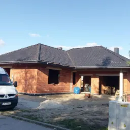 Nowo położony, grafitowy dach z dachówki ceramicznej na domu w trakcie budowy, z widoczną cegłą dziurawką, biały samochód dostawczy z otwartymi drzwiami i tablicą rejestracyjną ONA 7R82 zaparkowany...