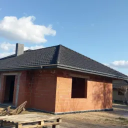 Surowy budynek jednorodzinny z ciemną dachówką, widoczne palety z materiałami budowlanymi i fragment sąsiedniego domu