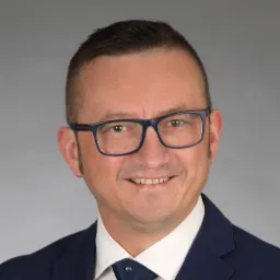 Portret uśmiechniętego mężczyzny w garniturze i okularach na szarym tle, z widocznym krawatem w drobne wzory.