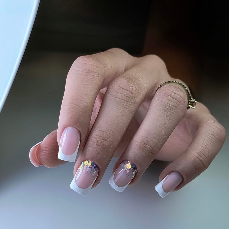 Dłoń z kwadratowymi paznokciami french manicure, dwa z brokatem. Pierścionek na palcu. Delikatne, naturalne tło. Precyzyjne wykonanie.