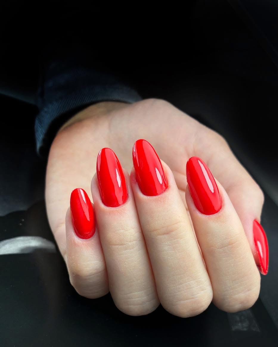 Dłoń z czerwonym, błyszczącym manicure hybrydowym na długich, owalnych paznokciach. Ciemne tło podkreśla intensywność koloru.