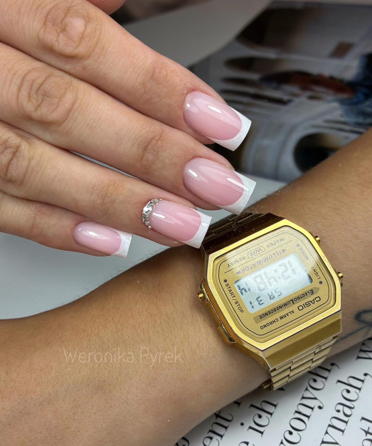 Dłoń z manicure francuskim w odcieniach różu i bieli, jeden paznokieć zdobiony cyrkoniami. Na nadgarstku złoty zegarek Casio. Subtelny i elegancki wygląd.