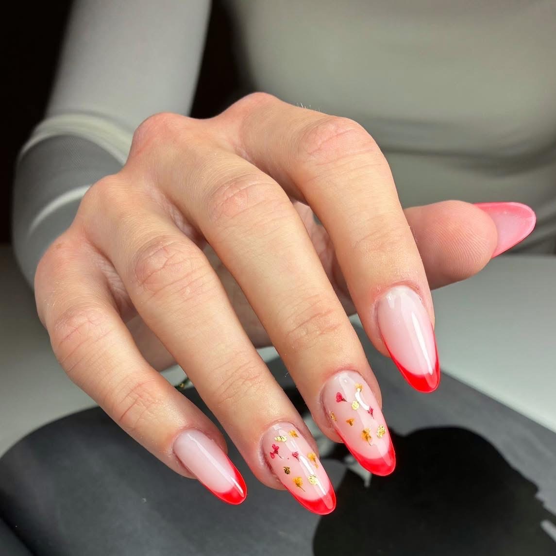Dłoń z czerwonym frenchem i drobnymi, złotymi zdobieniami w kształcie kwiatów na przezroczystej bazie. Migdałowy kształt paznokci, delikatne przejście kolorów.