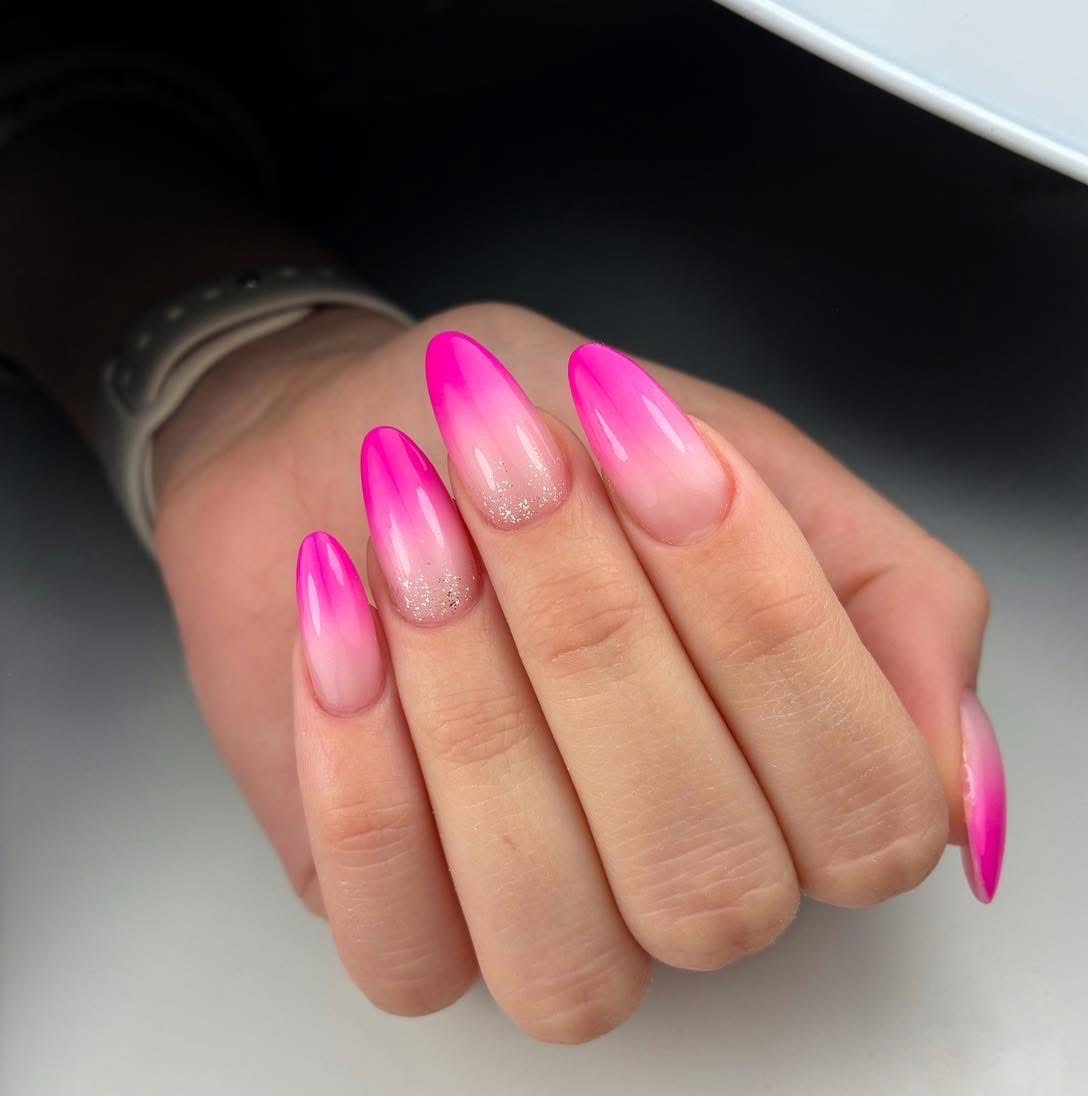 Dłoń z długimi, akrylowymi paznokciami w odcieniach różu z efektem ombre i delikatnym brokatem na serdecznym i środkowym palcu.