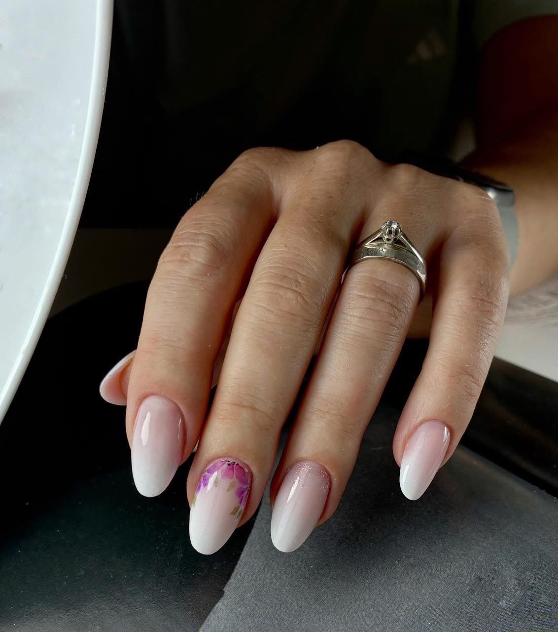 Dłoń z subtelnym manicure ombre, zdobiona kwiatowym wzorem na jednym paznokciu. Pierścionek na palcu dodaje elegancji. Delikatne przejście kolorów i precyzja wykonania.