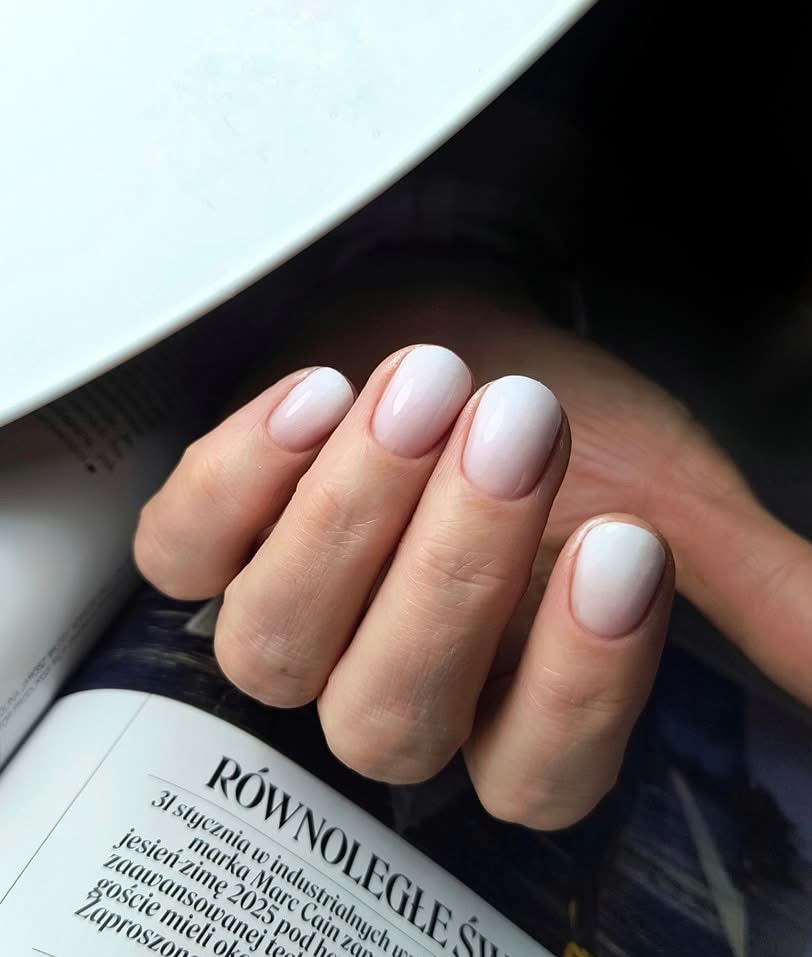 Delikatny manicure hybrydowy z efektem ombre w odcieniach bieli i różu, wykonany na naturalnych paznokciach o owalnym kształcie, na tle otwartego magazynu.