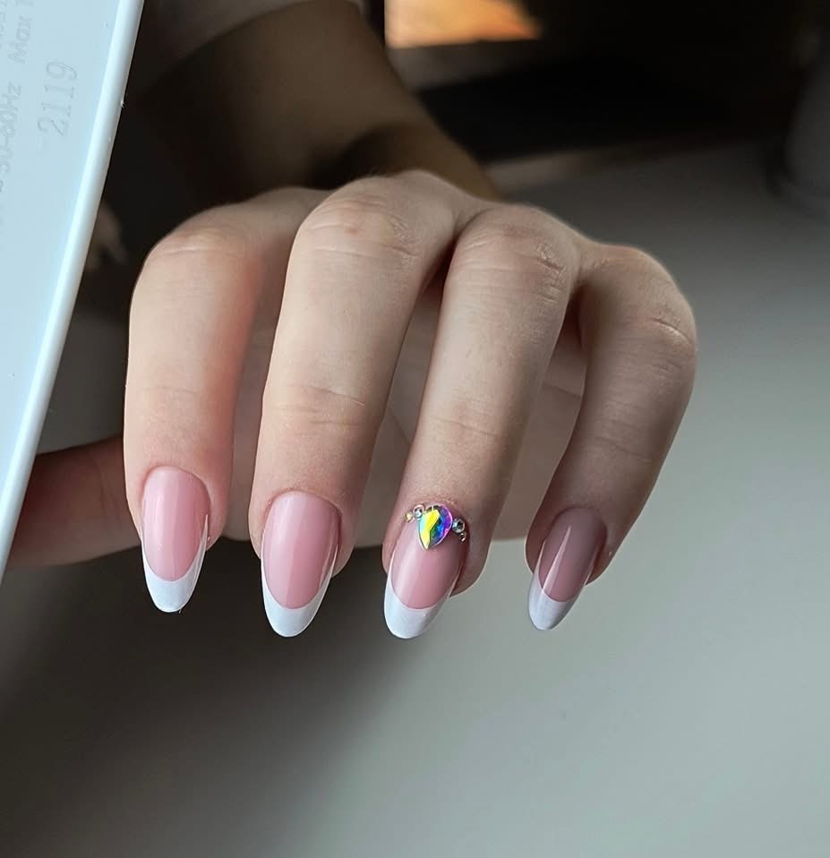 Dłoń z manicure francuskim, jeden paznokieć ozdobiony błyszczącym kamieniem w kształcie łzy. Delikatne, jasne tło. Precyzyjne wykonanie.