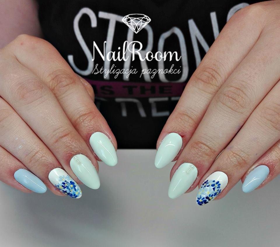 Dłonie z pastelowymi paznokciami w odcieniach błękitu i mięty, jeden paznokieć ozdobiony drobnymi niebieskimi kwiatkami, w tle logo salonu stylizacji paznokci.