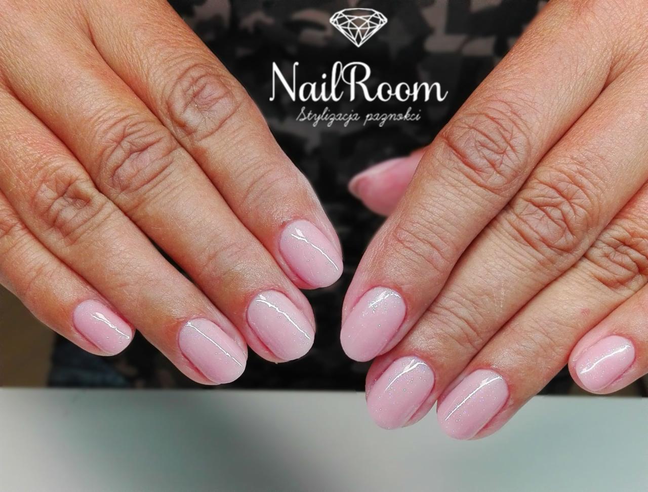 Dłonie z delikatnym, różowym manicure hybrydowym, ozdobionym cienką, srebrną linią i subtelnym brokatem, na tle z logo salonu.