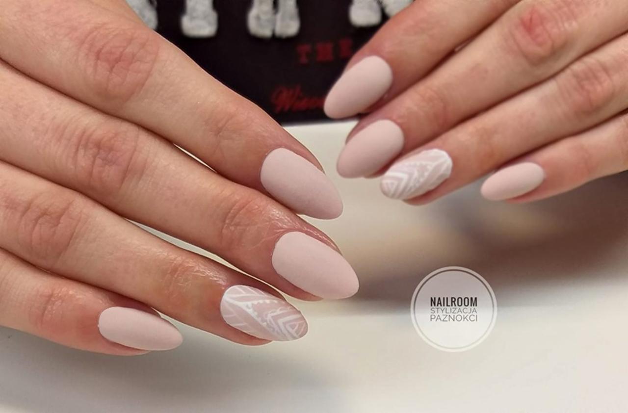 Delikatne, matowe paznokcie w odcieniach beżu z białym, geometrycznym wzorem na dwóch palcach, prezentowane na dłoniach na jasnym tle.