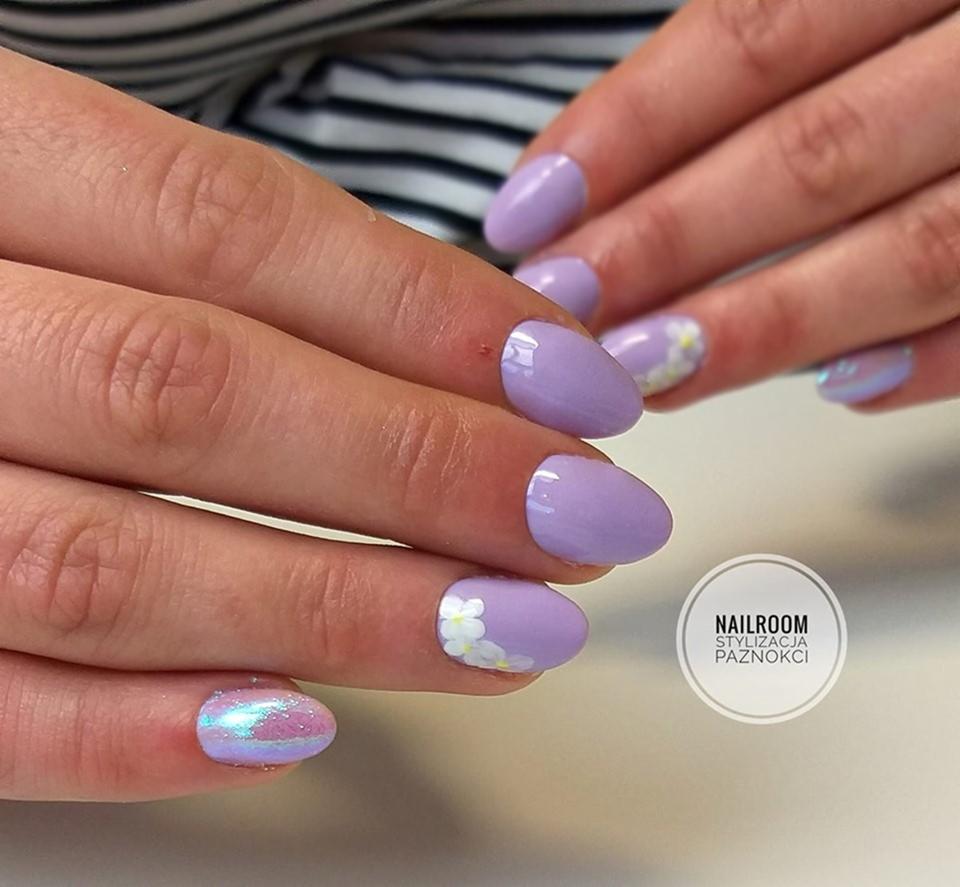 Dłonie z owalnymi paznokciami pomalowanymi na liliowo, z delikatnym wzorem białych kwiatków na niektórych paznokciach i opalizującym efektem na jednym, na tle w paski.