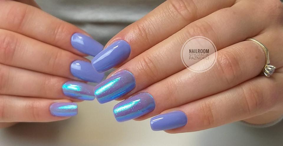 Dłonie z pomalowanymi na fioletowo paznokciami, niektóre z efektem opalizującym na niebiesko-zielono, pierścionek z oczkiem na palcu.