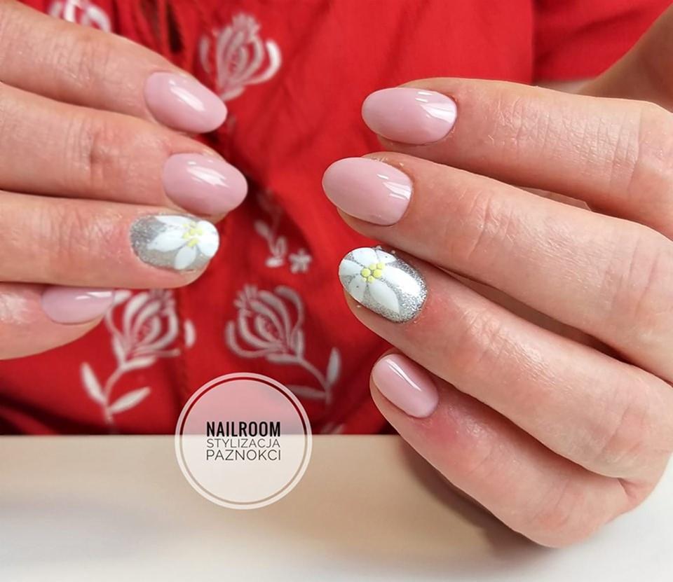 Delikatny, pudrowy manicure z akcentem srebrnego brokatu i białych kwiatków na dwóch paznokciach, na tle czerwonej bluzki z kwiatowym wzorem.
