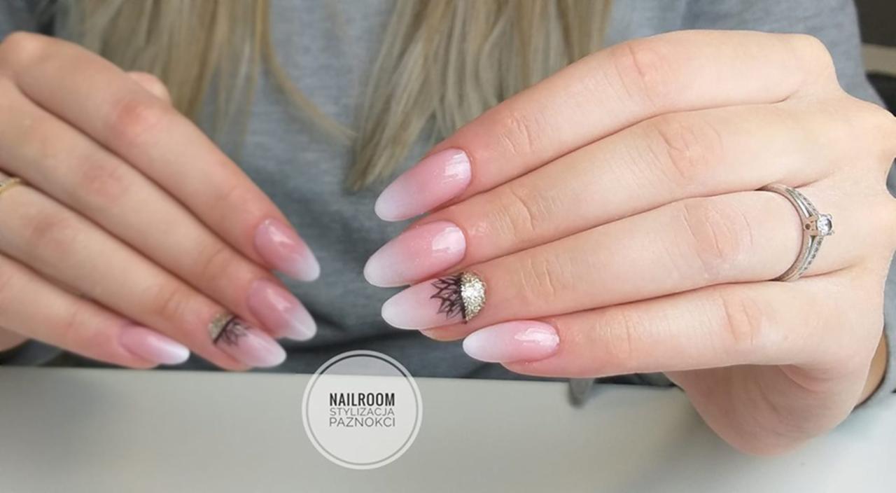 Dłonie z długimi, ombre paznokciami w odcieniach różu i bieli, zdobionymi drobnymi, złotymi elementami i czarnym wzorem przypominającym koronę, z pierścionkiem na palcu serdecznym.