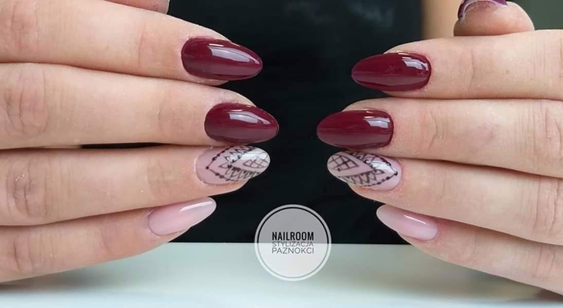 Dłonie z manicure hybrydowym: trzy paznokcie w odcieniach bordo, jeden jasnoróżowy i jeden jasnoróżowy z czarnym, geometrycznym wzorem koronki.