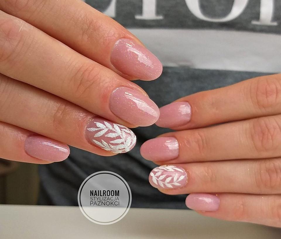 Delikatne, różowe paznokcie z białym, ręcznie malowanym wzorem liści na dwóch palcach, prezentowane na tle szarej koszulki.