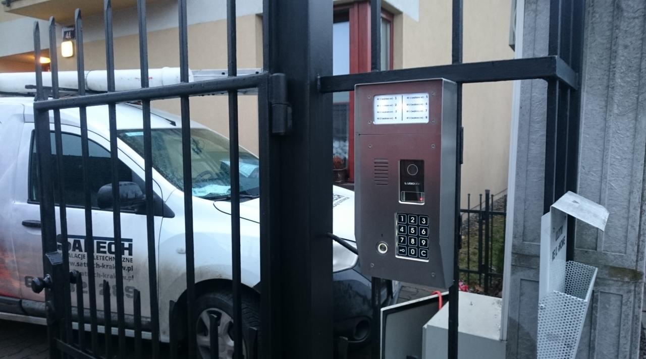 Nowoczesny domofon z klawiaturą numeryczną zamontowany na czarnej, metalowej bramie wejściowej, z widocznym samochodem serwisowym firmy instalacyjnej w tle.