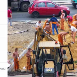 Przygotowanie terenu pod kostkę brukową: grupa robotników w pomarańczowych kombinezonach rozgarnia piasek łopatami, w tle zaparkowane samochody i koparko-ładowarka CAT.