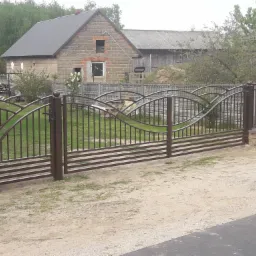 Brązowa brama wjazdowa z metalowymi prętami i dekoracyjnymi łukami, osadzona na niskim murku z cegły, na tle wiejskiego domu z cegły i drewnianej stodoły.