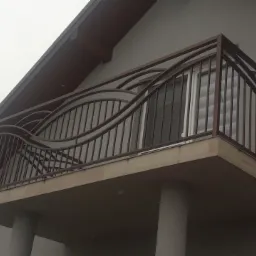 Balkon z metalową balustradą o falistym wzorze, brązowa konstrukcja na tle szarej elewacji budynku.