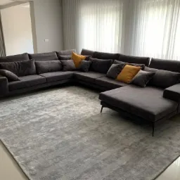 Duży, narożny, ciemnoszary tapicerowany zestaw wypoczynkowy z poduszkami w odcieniach szarości i musztardowej żółci, ustawiony na wzorzystym, srebrnym dywanie w przestronnym salonie z jasnymi...