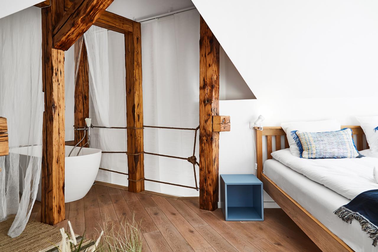 Jasne wnętrze pokoju z drewnianymi belkami, białą wanną za zasłoną, łóżkiem z drewnianą ramą i niebieskim stolikiem nocnym.