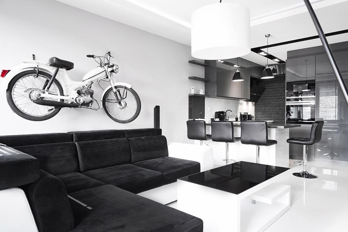 Nowoczesne wnętrze w czerni i bieli z motocyklem na ścianie, widok na otwartą kuchnię z barkiem i czarnymi hokerami, biała podłoga z połyskiem.