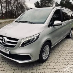 Srebrny Mercedes-Benz V-klasy na parkingu z kostki, znak drogowy P w tle. Auto z relingami dachowymi i przyciemnianymi szybami.