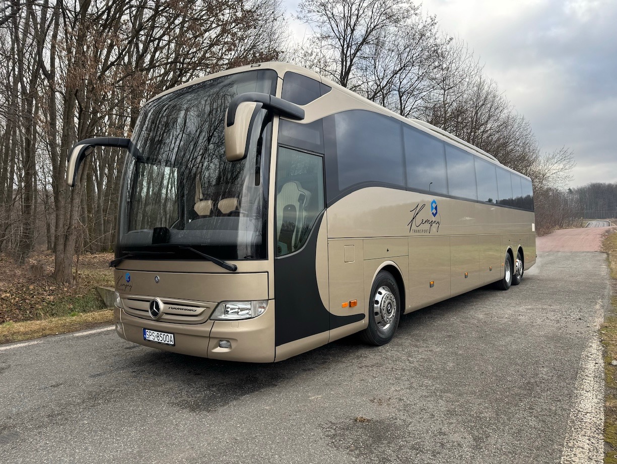 Złoty autokar Mercedes-Benz Tourismo z czarnymi akcentami, zaparkowany na drodze w pobliżu lasu. Widoczny znak firmowy i tablica rejestracyjna.