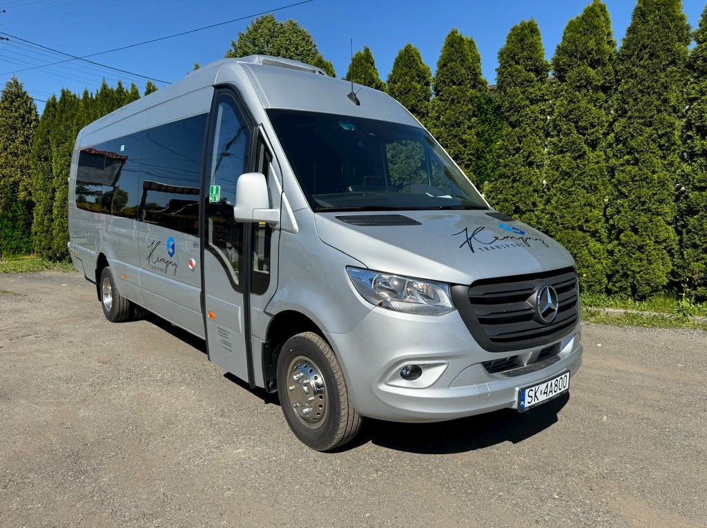 Srebrny bus Mercedes-Benz z logo firmy transportowej na tle zielonych drzew, zaparkowany na szutrowej nawierzchni w słoneczny dzień.