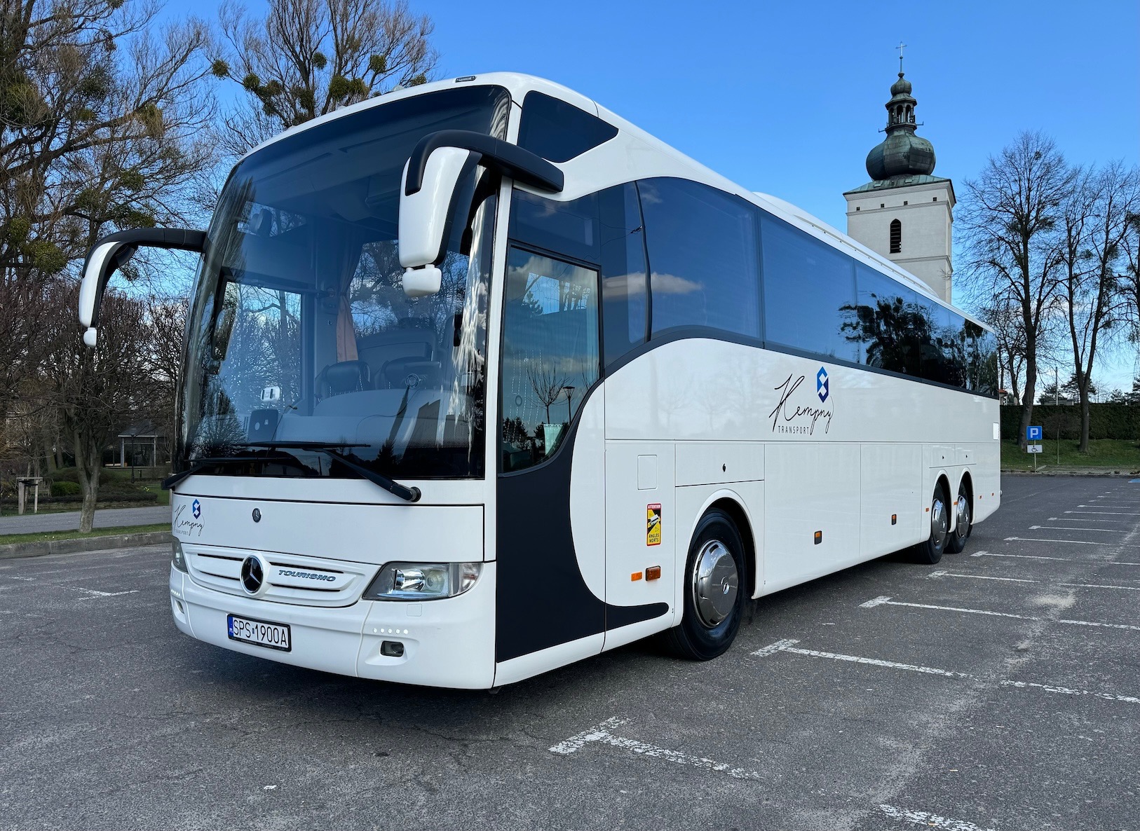 Nowoczesny, biały autokar z logo firmy na parkingu, w tle wieża kościoła. Autokar marki Mercedes-Benz Tourismo z czarnymi akcentami i dużymi oknami.