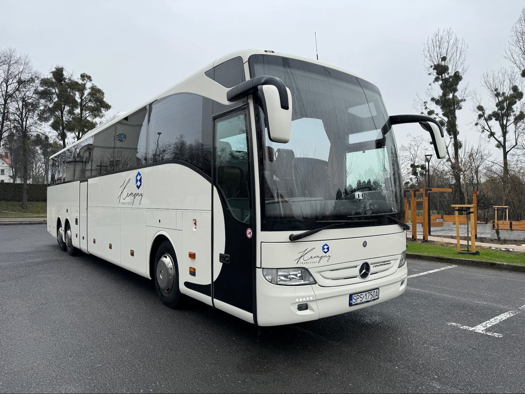 Biały autokar turystyczny Mercedes-Benz z logo firmy na parkingu, widok z przodu pod pochmurnym niebem. Widoczne drzewa i elementy małej architektury w tle.