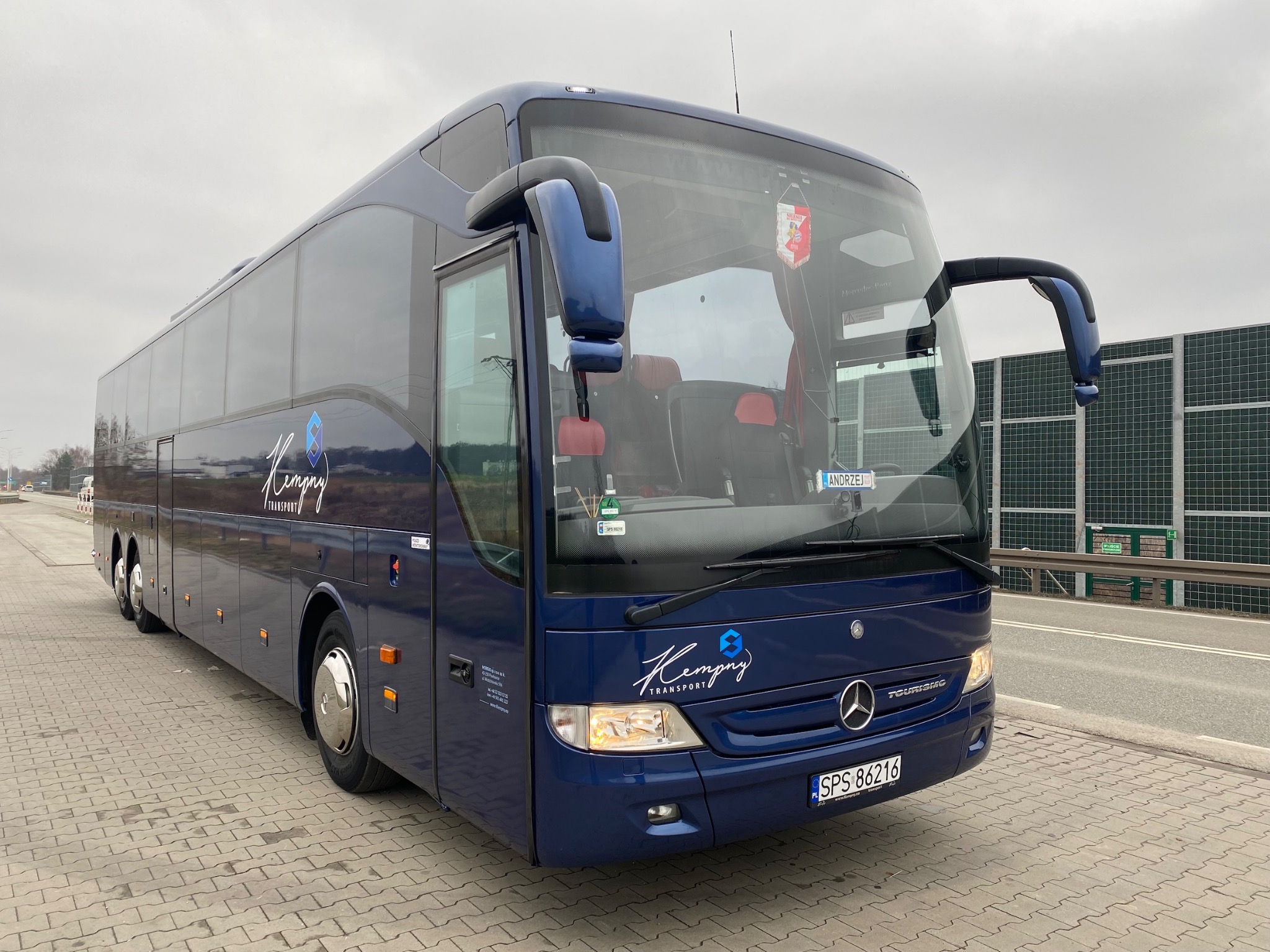 Niebieski autokar marki Mercedes-Benz Tourismo z logo firmy Kempny Transport, zaparkowany na chodniku z kostki brukowej. Widok z przodu i z boku, tablica rejestracyjna SPS 86216.