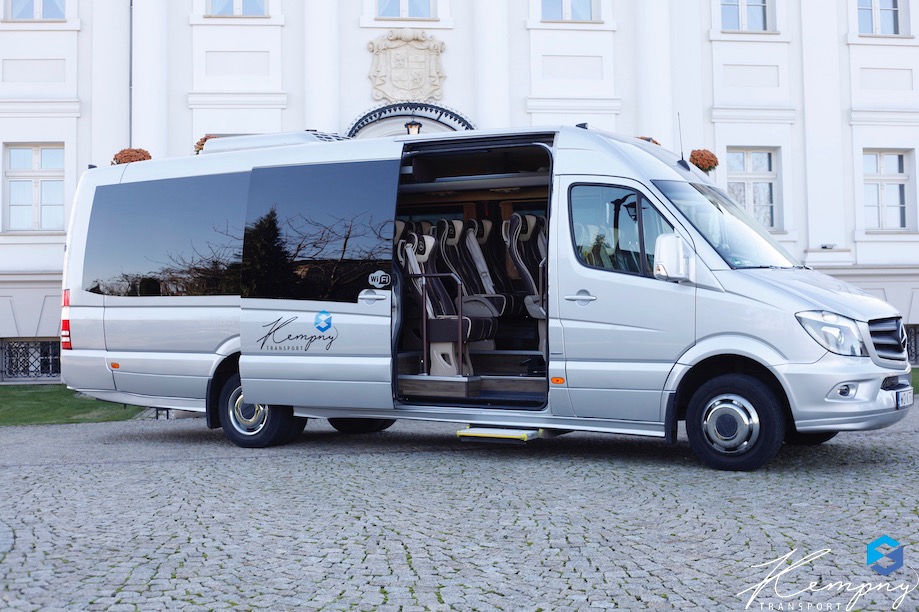 Srebrny bus z otwartymi drzwiami na tle jasnego budynku. Widoczne fotele pasażerskie. Logo firmy na karoserii. Kostka brukowa na pierwszym planie.