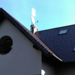 Widok z dołu na fragment domu z ciemnobrązowym dachem z dachówką bitumiczną, dwoma oknami dachowymi, okrągłym oknem na szczycie i anteną satelitarną na kominie.