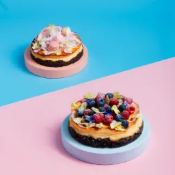 Dwa mini serniki baskijskie na pastelowych podstawkach, jeden z makaronikami i bezami, drugi z malinami i jagodami, na tle w kolorach różu i błękitu.